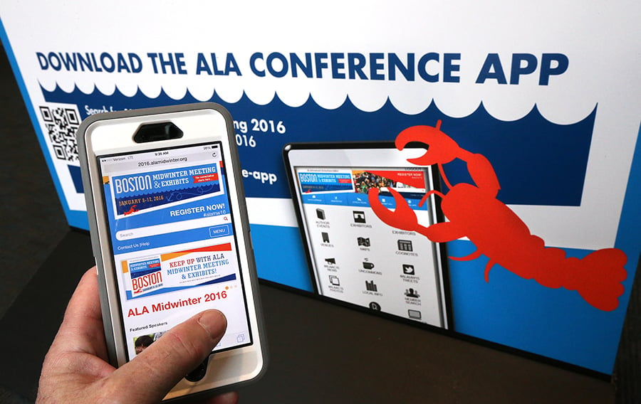 An ALA Midwinter 2016 attendee holds an iPhone with the ALA Midwinter app in front of a sign that reads Download the Midwinter App.