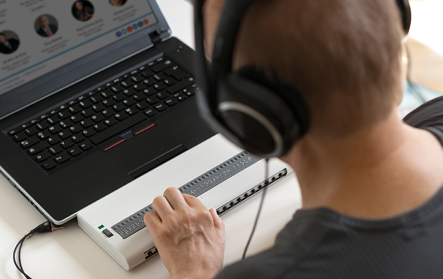 A man in a gray t-shirt with headphones on uses a braille keyboard to navigate an eventScribe website on his laptop.